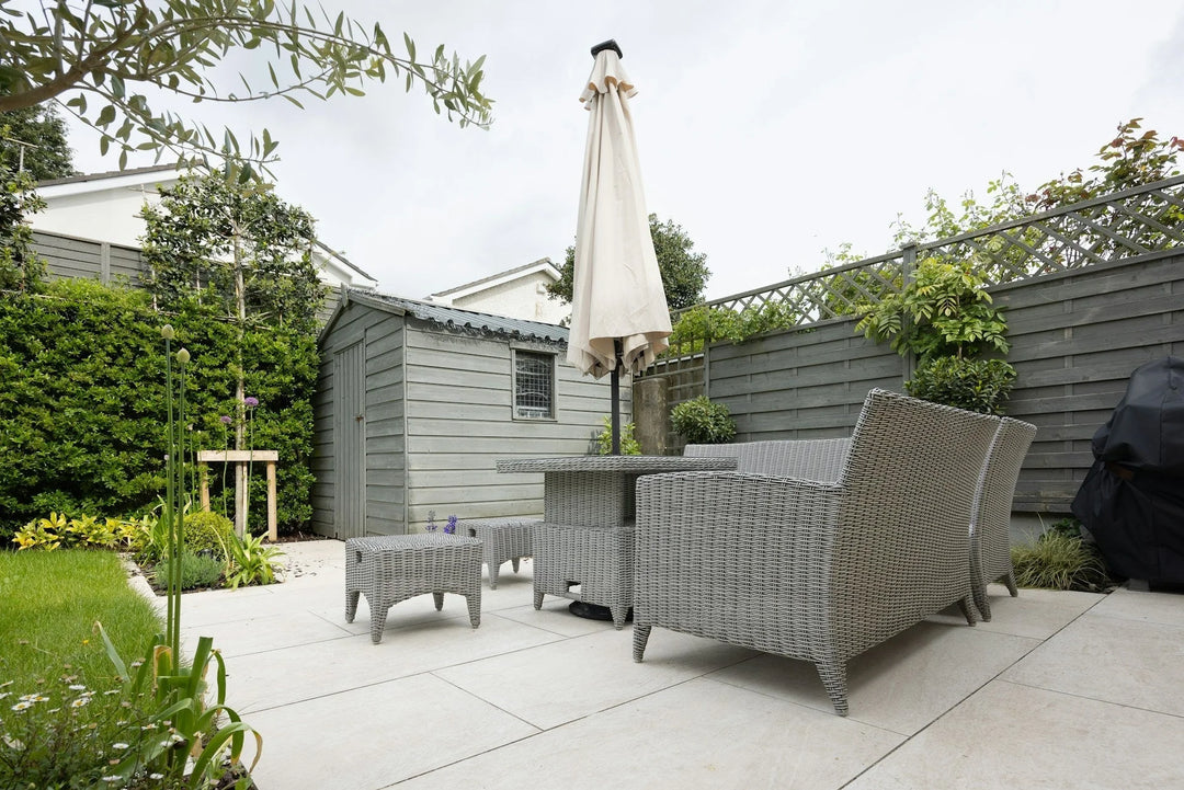 Gray wicker patio furniture with an umbrella, surrounded by greenery in a garden. Refresh Your Patio & Garden This Summer.