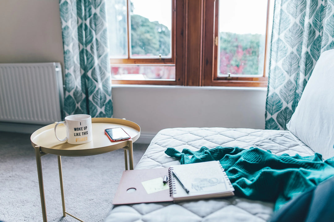 A cozy seating area with a gold side table, a white mug, a smartphone, a sketchbook, and a green blanket. Grieving in Isolation: Surviving the Process in COVID.