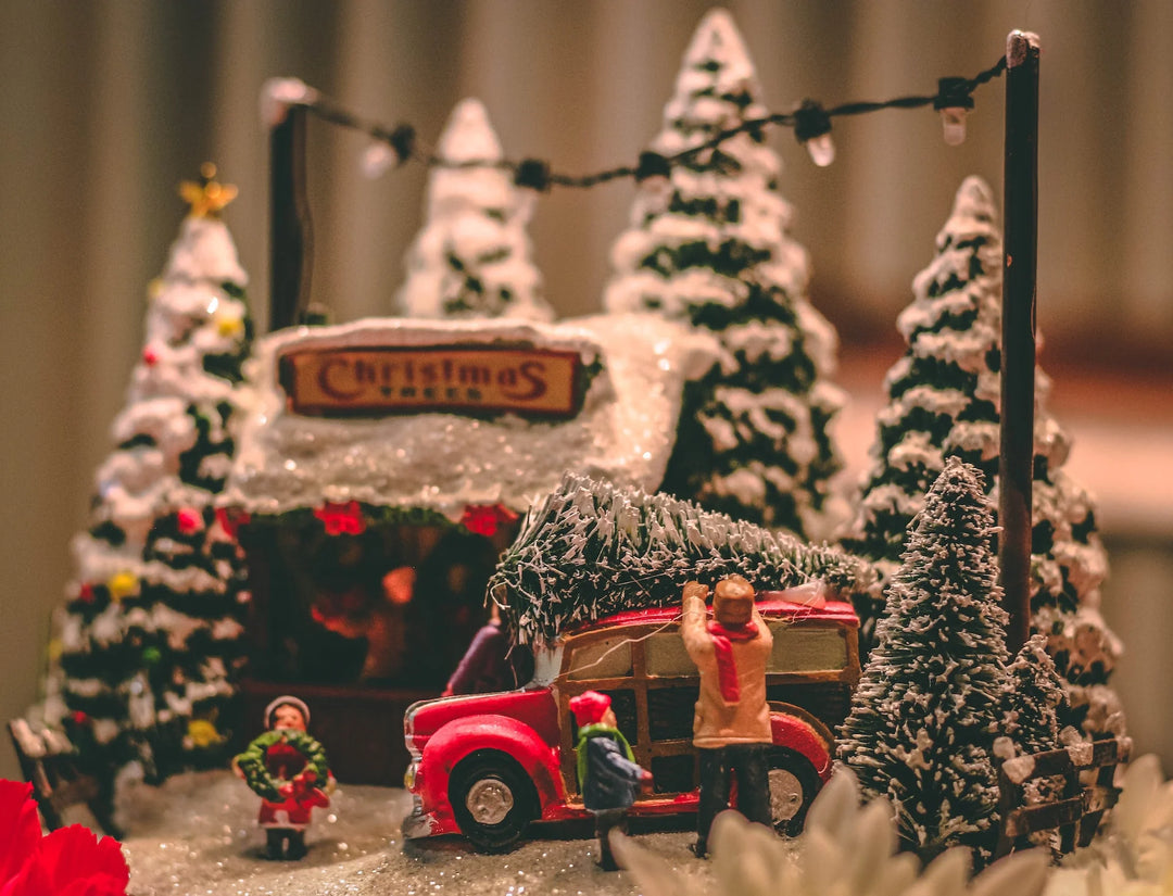 A festive miniature scene with a red truck carrying a Christmas tree, snow-covered trees, and a 'Christmas Town' sign.