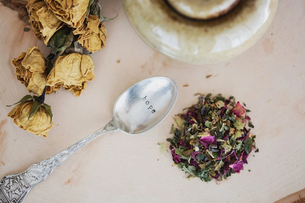 Herbal blend with dried rose petals on wood, silver spoon engraved 'hope', and dried flowers nearby. Self-Care and Grieving: Gift Ideas to Nourish the Soul.