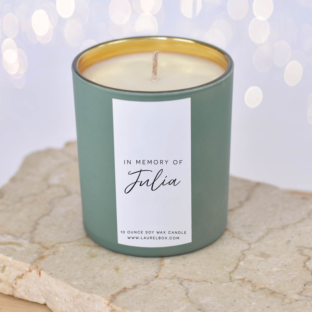 A close-up of a light green candle with the text "In Memory of Julia" on a white label sitting on a textured surface with blurred lights in the background.