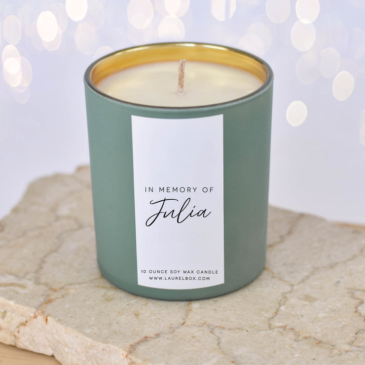 A close-up of a light green candle with the text "In Memory of Julia" on a white label sitting on a textured surface with blurred lights in the background.