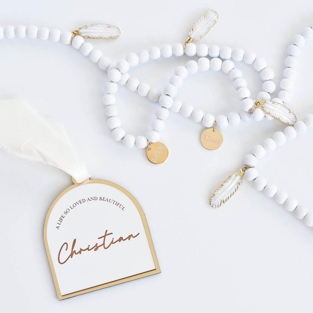 A close-up of white and gold ornament on the left with the text "A Life So Loved And Beautiful, Christian" with a white silk ribbon, and at the top, a white garland with white feather charms and engraved gold charms against a white background.