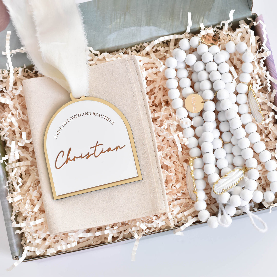 A close-up of a laurelbox gift set with a white and gold ornament on the left with the text "A Life So Loved And Beautiful, Christian" with a white silk ribbon, and at the top, a white garland with white feather charms and engraved gold charms.