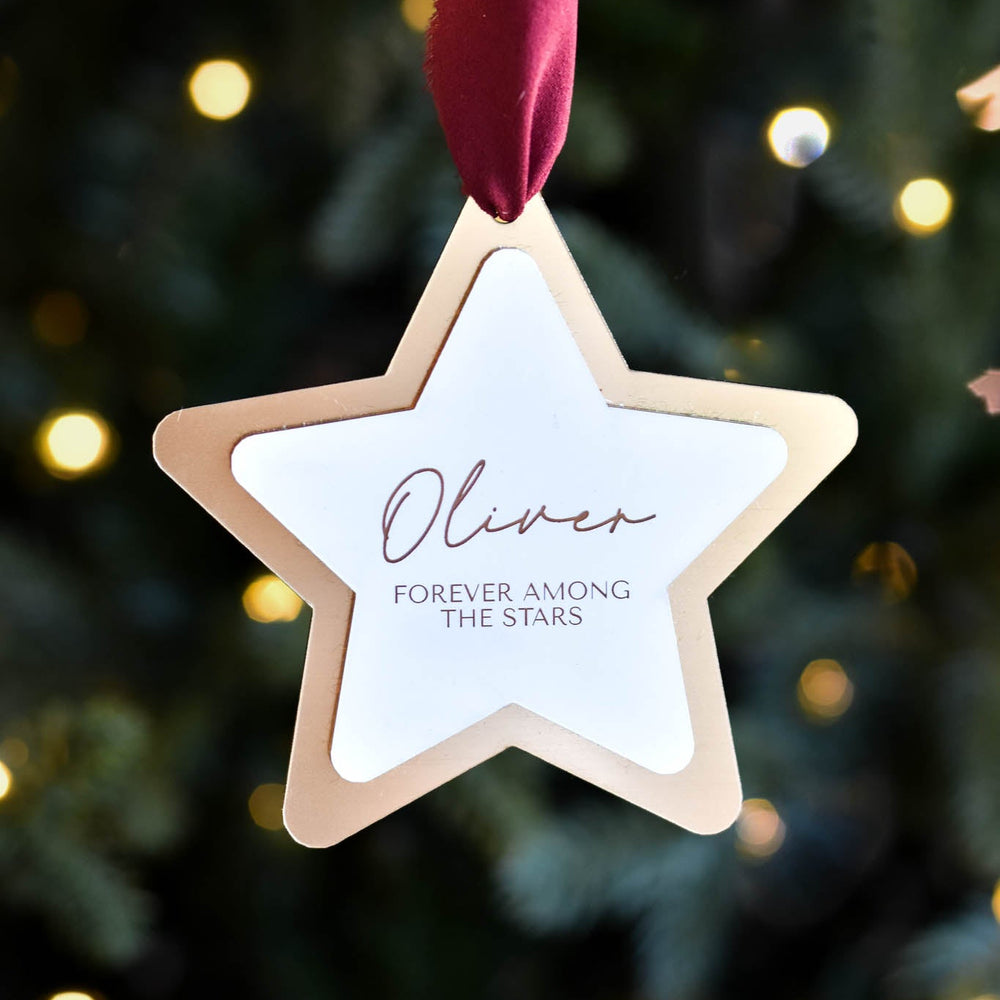 Layered star ornament in white and gold with the name 'Oliver' and text 'FOREVER AMONG THE STARS', hung with a maroon ribbon.