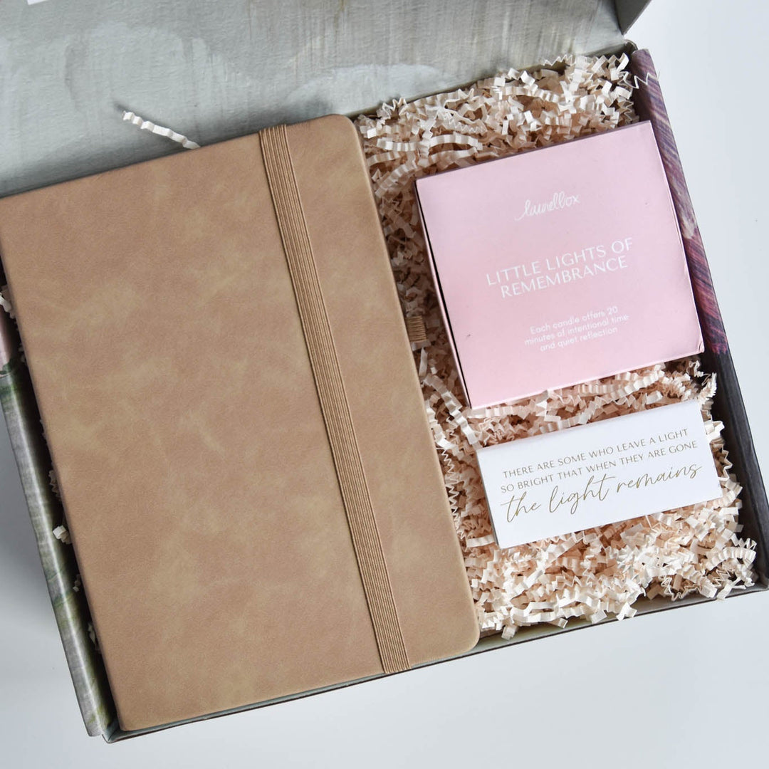 A gift box from laurelbox containing a tan leather notebook, a pink candle set reading 'Little Lights of Remembering', and a white matches set on shredded paper.