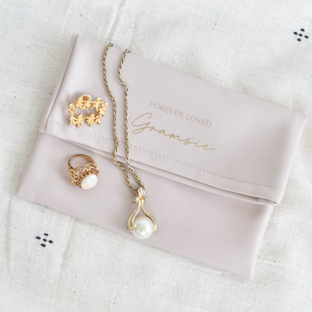 A gold-toned jewelry set including a pendant necklace with a pearl drop, a ring with a pearl and gold crown motif, and a small gold charm, arranged on a pale pink clutch bag with the words 'FOREVER LOVED Gramsie' printed on it, all placed on a white textured fabric background.