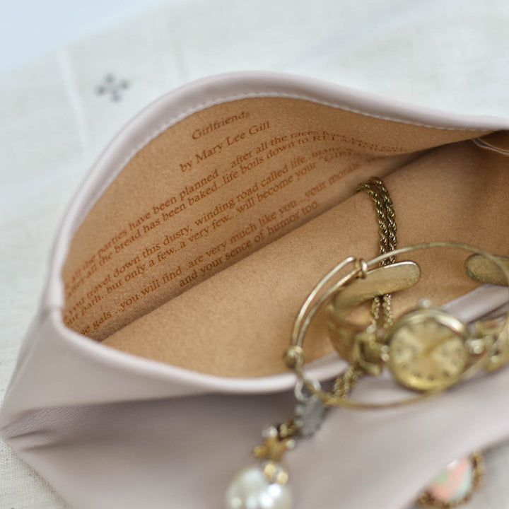 A blush pink fabric jewelry pouch with a printed book-style interior, shown open with gold-toned jewelry inside on a light fabric surface.