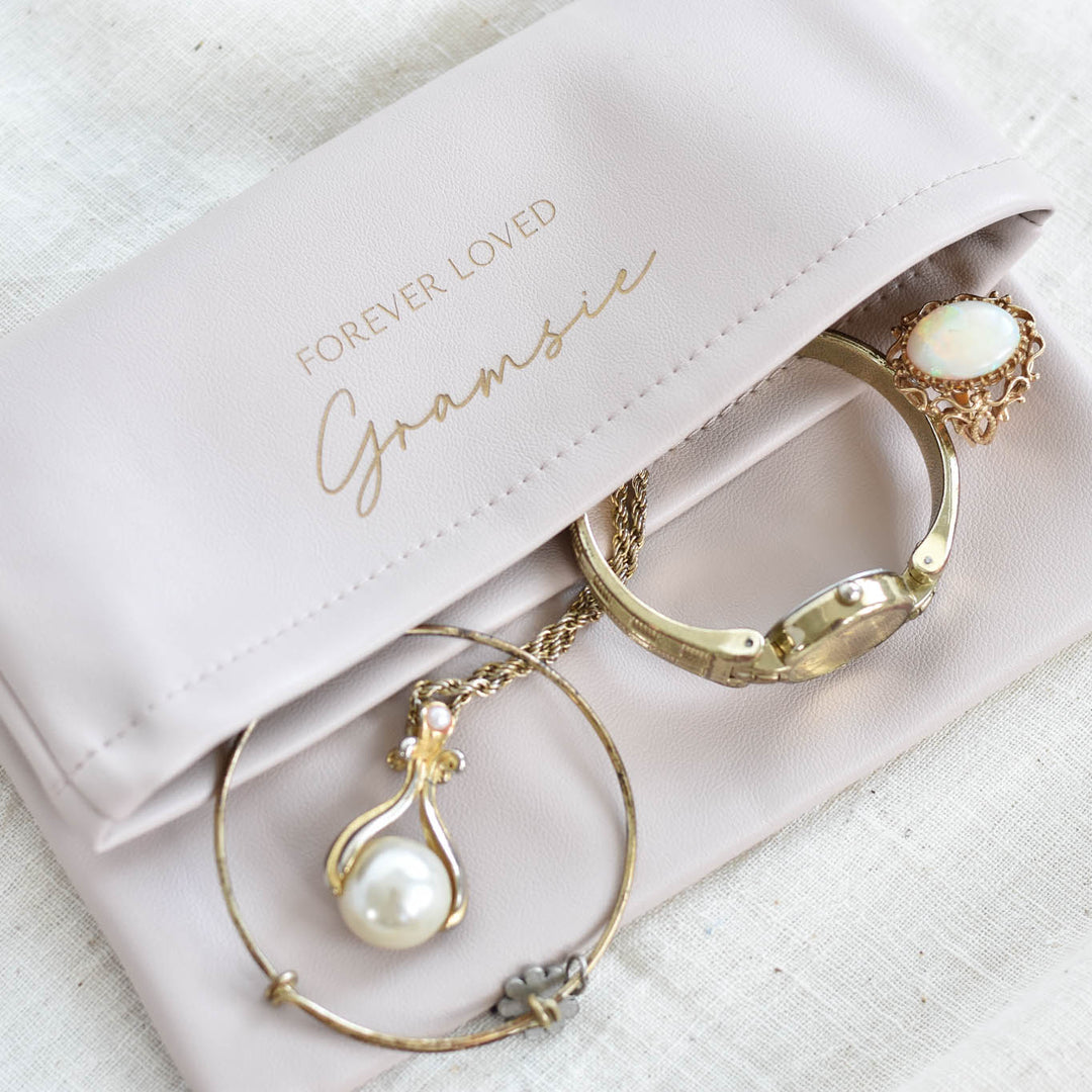 A pale blush pink satin jewelry pouch with gold foil lettering that reads 'FOREVER LOVED Gramsie', styled with gold jewelry on a textured white linen background.
