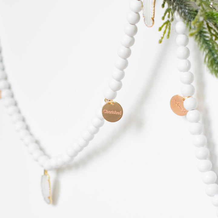 A close-up of a white garland with white feather charms and engraved gold charms highlighting the cherished charm and feather charm against a white background and pine branches.