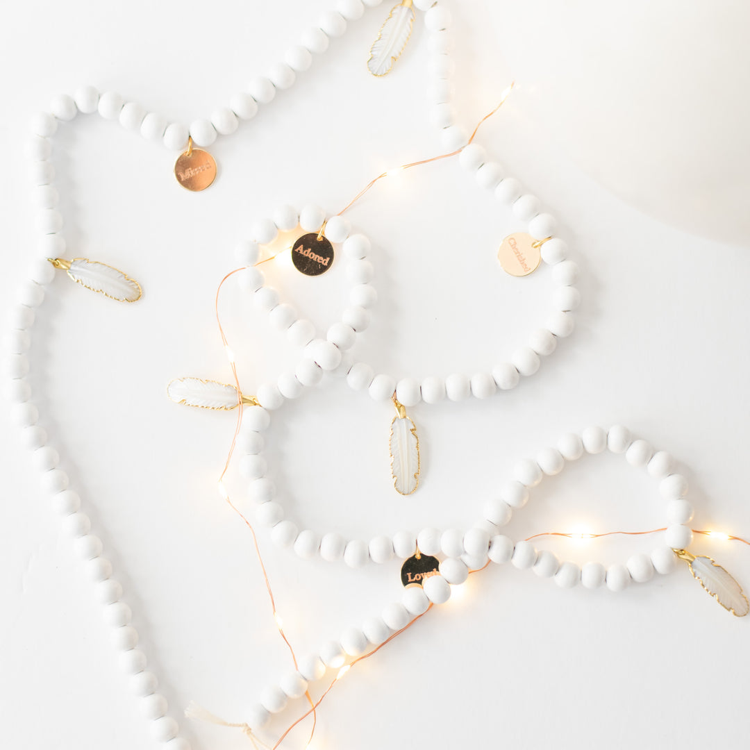A close-up of a white garland with white feather charms and engraved gold charms, highlighting the gold charms with the "Missed", "Adorned", and "Loved" text with lights scattered around the white background.