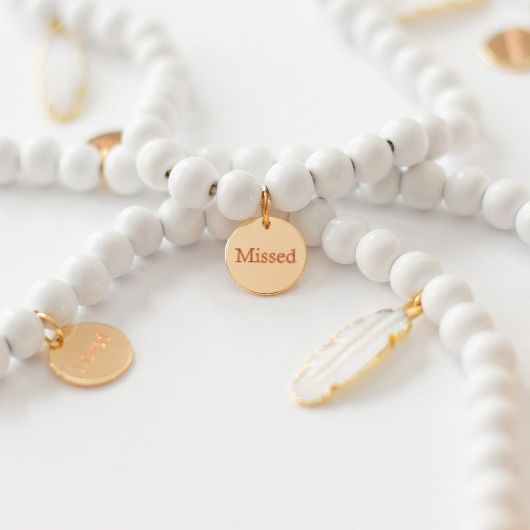 A close-up of a white garland with white feather charms and engraved gold charms, highlighting the gold charm with the "Missed" text.