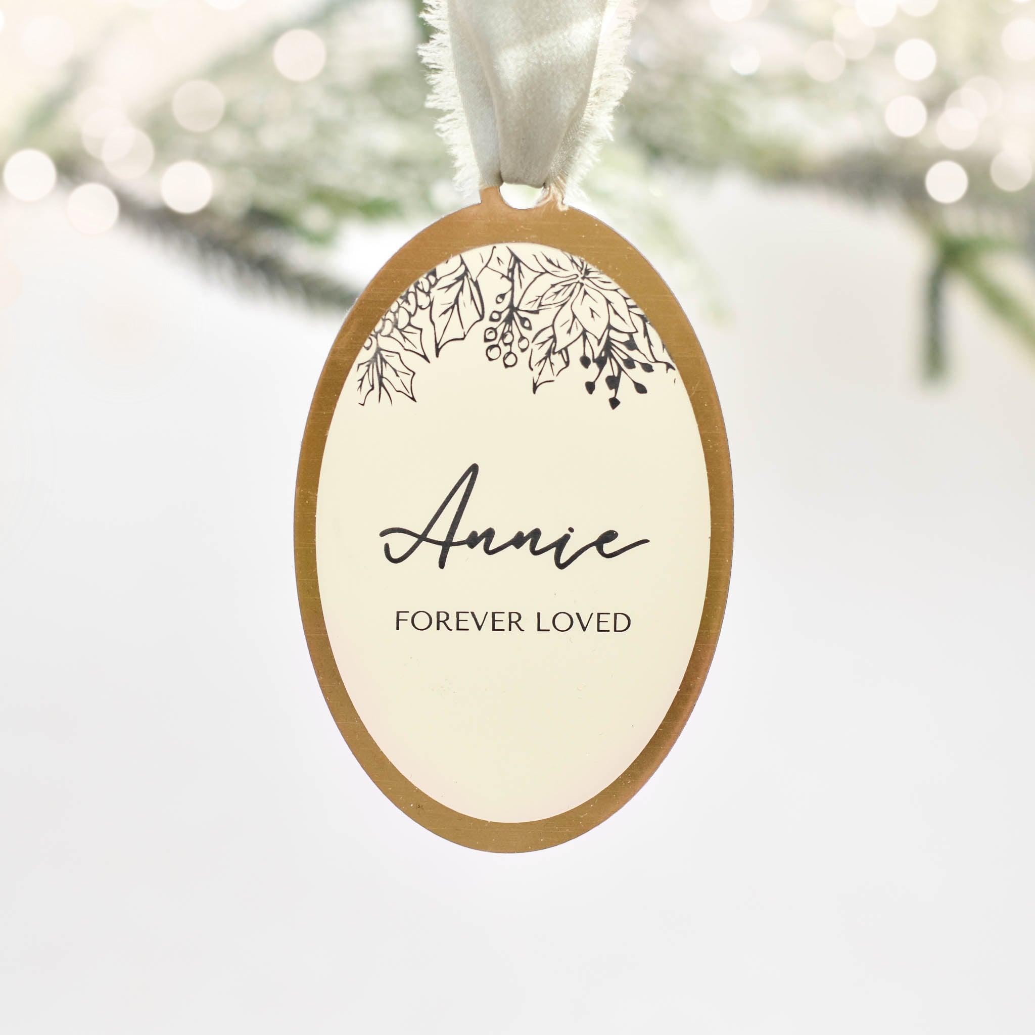 A close-up of a cream and gold oval-shaped ornament with white silk ribbon at the top, with 'Annie Forever Loved' text in the middle against a white background.