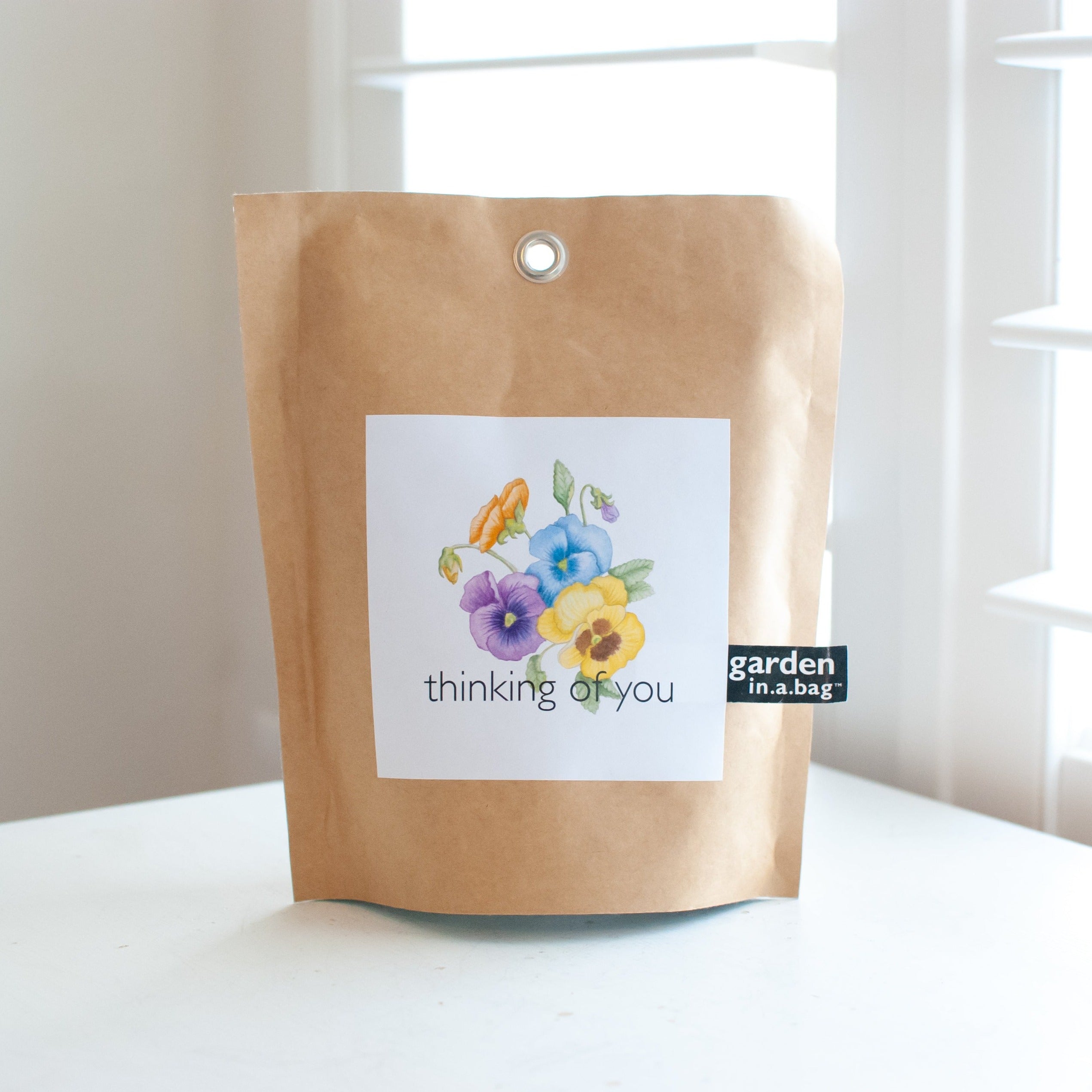 A close-up of a brown bag with a Thinking of You sticker with flowers and garden in a bag text on the right sitting on a white table