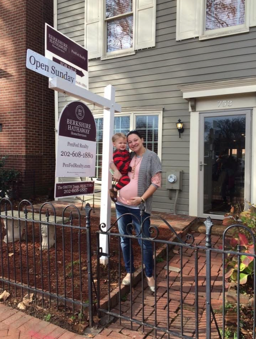 A woman who is pregnant standing in front of a townhome with a for sale sign in front of it, holding a toddler. 