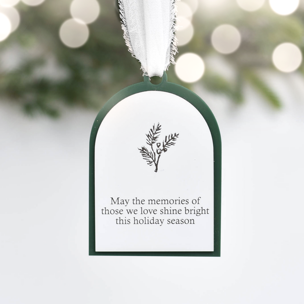 A white arched hanging ornament with a dark green border, featuring a small holly illustration and the text 'May the memories of those we love shine bright this holiday season.' It hangs from a white ribbon against a softly blurred holiday background with lights and greenery.