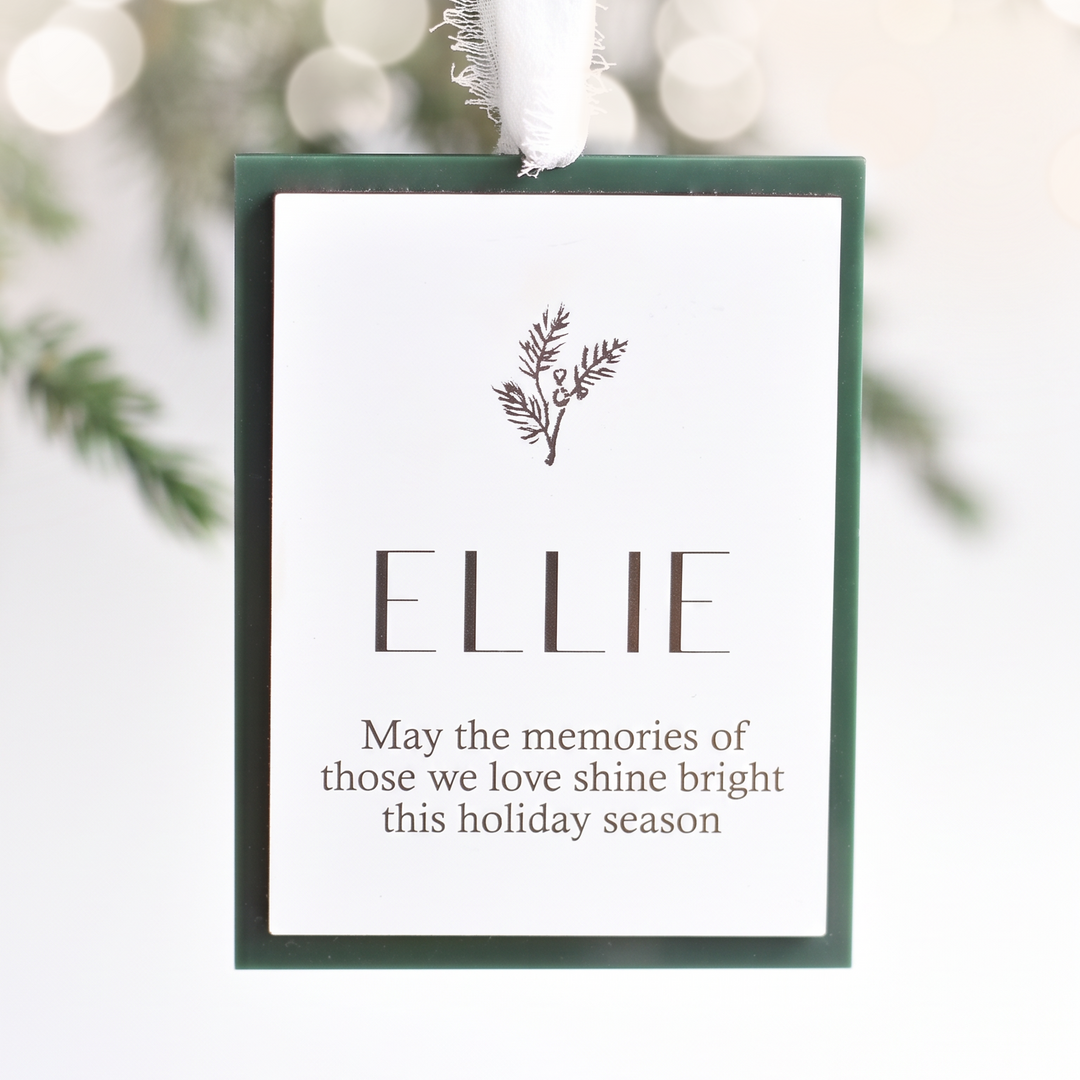 A personalized Christmas ornament tag with a white background, dark green border, and a white hanging ribbon. The tag is personalized with the name 'ELLIE' and a small sprig illustration, set against a soft, blurred holiday background.