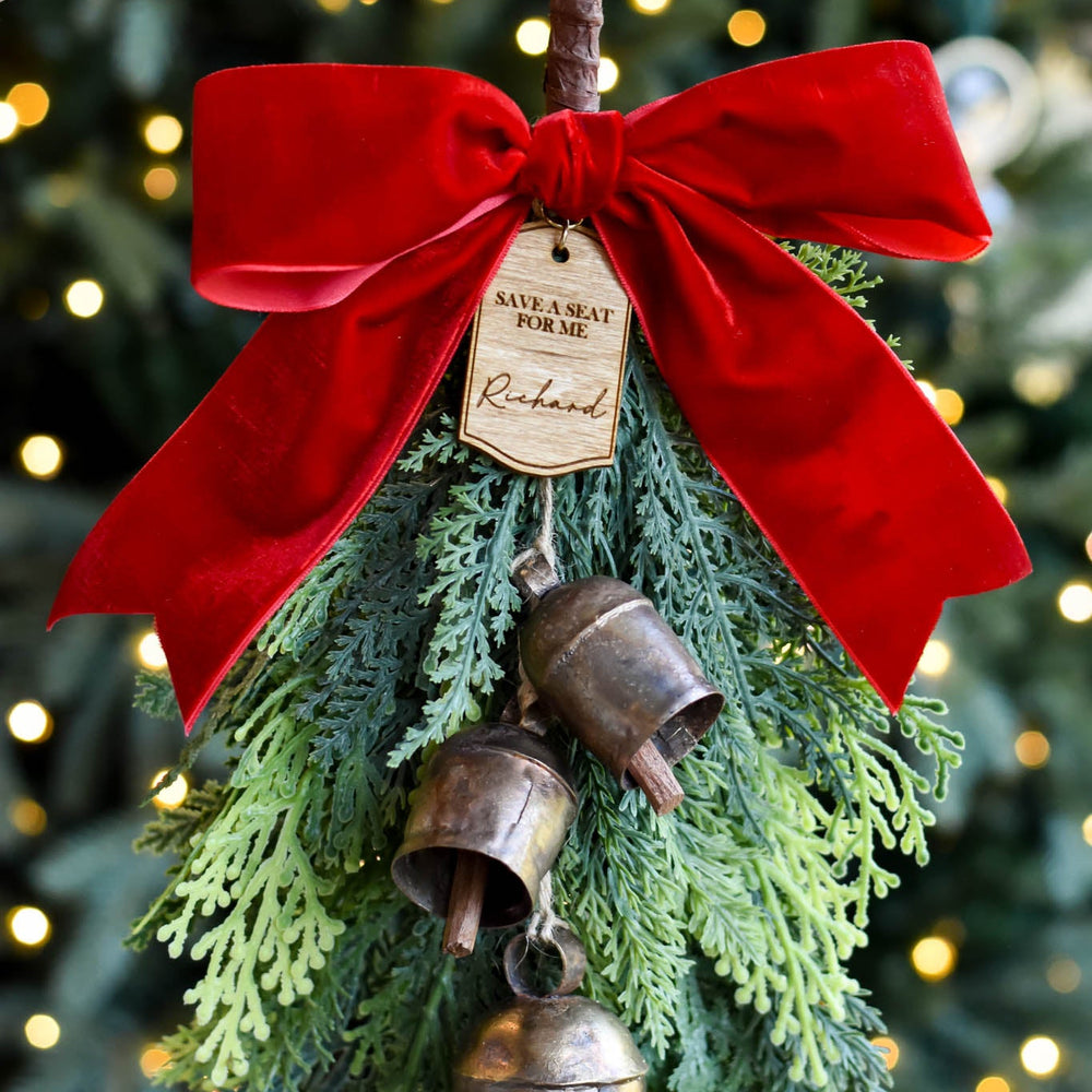 A hand holds a rustic Christmas ornament featuring a large red velvet bow, green pine branches, hanging bronze bells, and a small wooden tag, with a blurred Christmas tree in the background.
