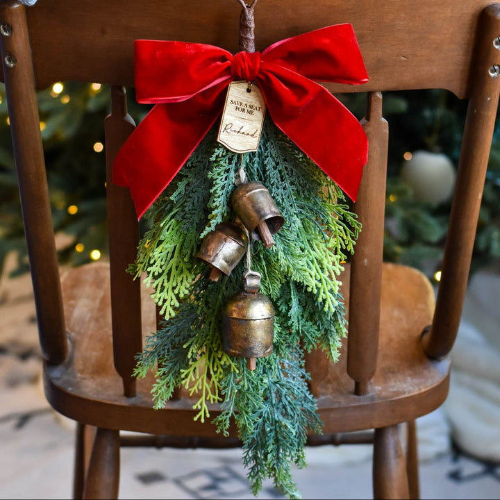 A hand holds a small Christmas ornament shaped like a miniature evergreen tree with a red velvet bow, brass bells, and a hanging tag, displayed on a wooden chair in front of a blurred Christmas tree.