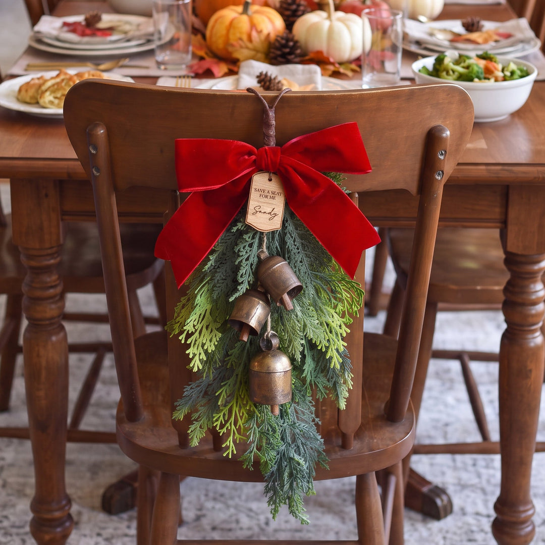 A holiday greenery evergreen swag with a red velvet bow, brass bells, and a hanging tag, displayed on a wooden chair in front of a Thanksgiving table.