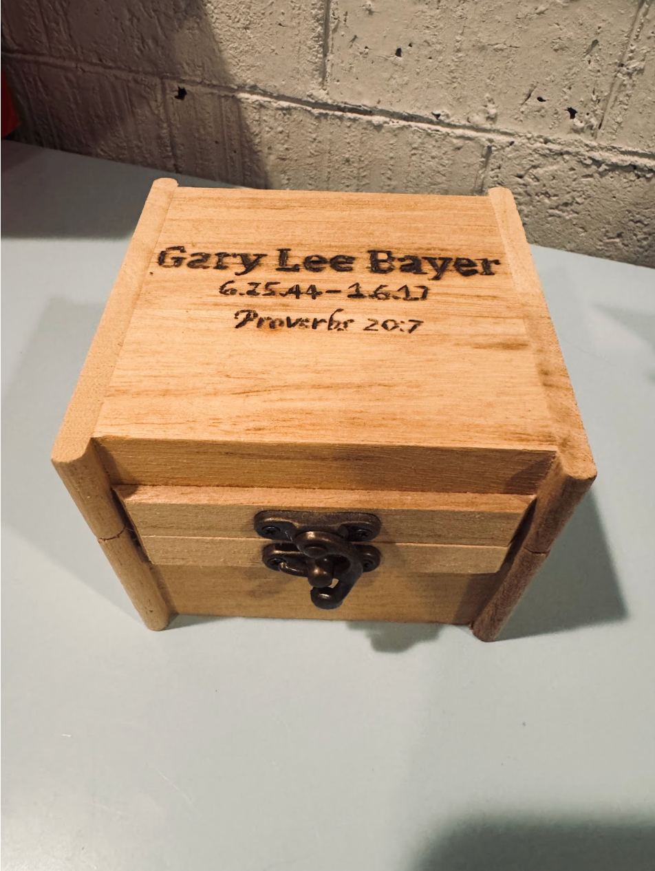 closed wooden box with the words "Gary Lee Bayer 6.25.44-1.6.17 Proverbs 20:7" branded on the top
