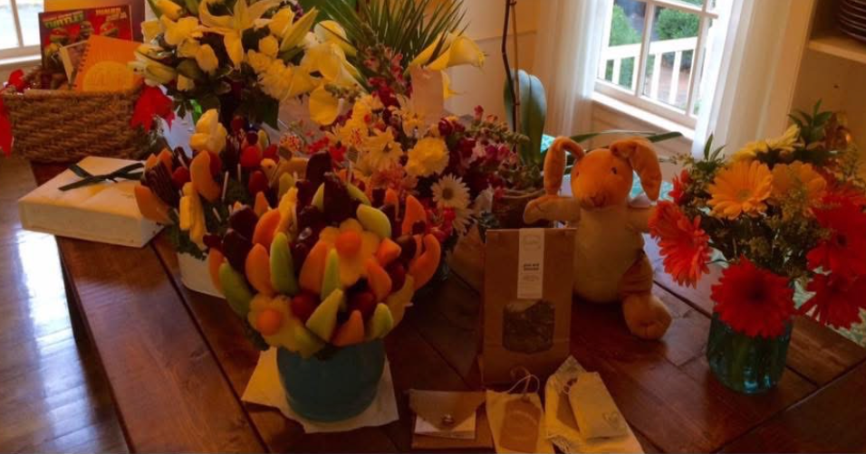 photo of edible arrangements, vases of flowers, cards, stuffed animals and baskets sitting on a wooden table