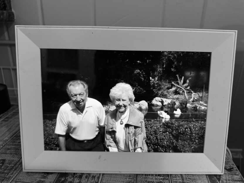 Black and white photo of an elderly couple outdoors, smiling, with a pond and ducks in the background, framed in white.