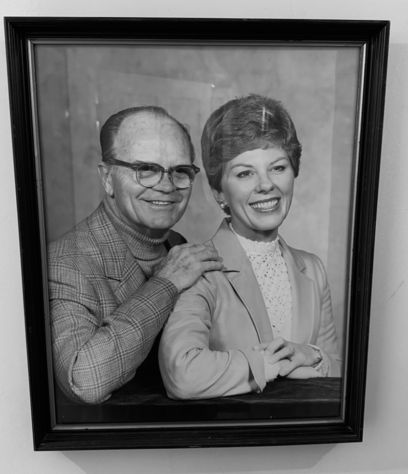 Black and white portrait of an older couple, man with glasses in a plaid suit, woman with short hair in a cream jacket, both smiling.
