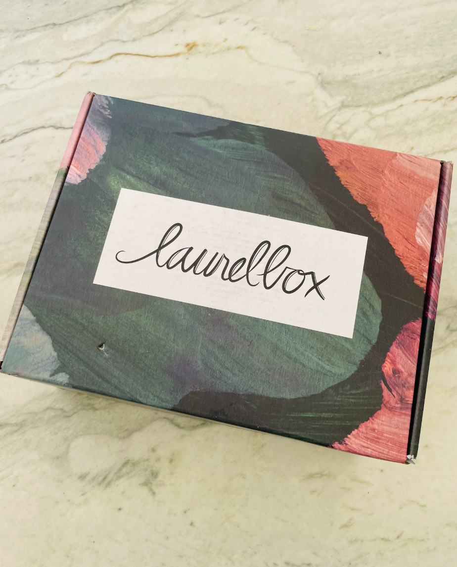 A rectangular product box with a colorful abstract painted design in green, black, and coral pink, featuring a white label with the script brand name 'laurelbox' on top, placed on a light marble surface.