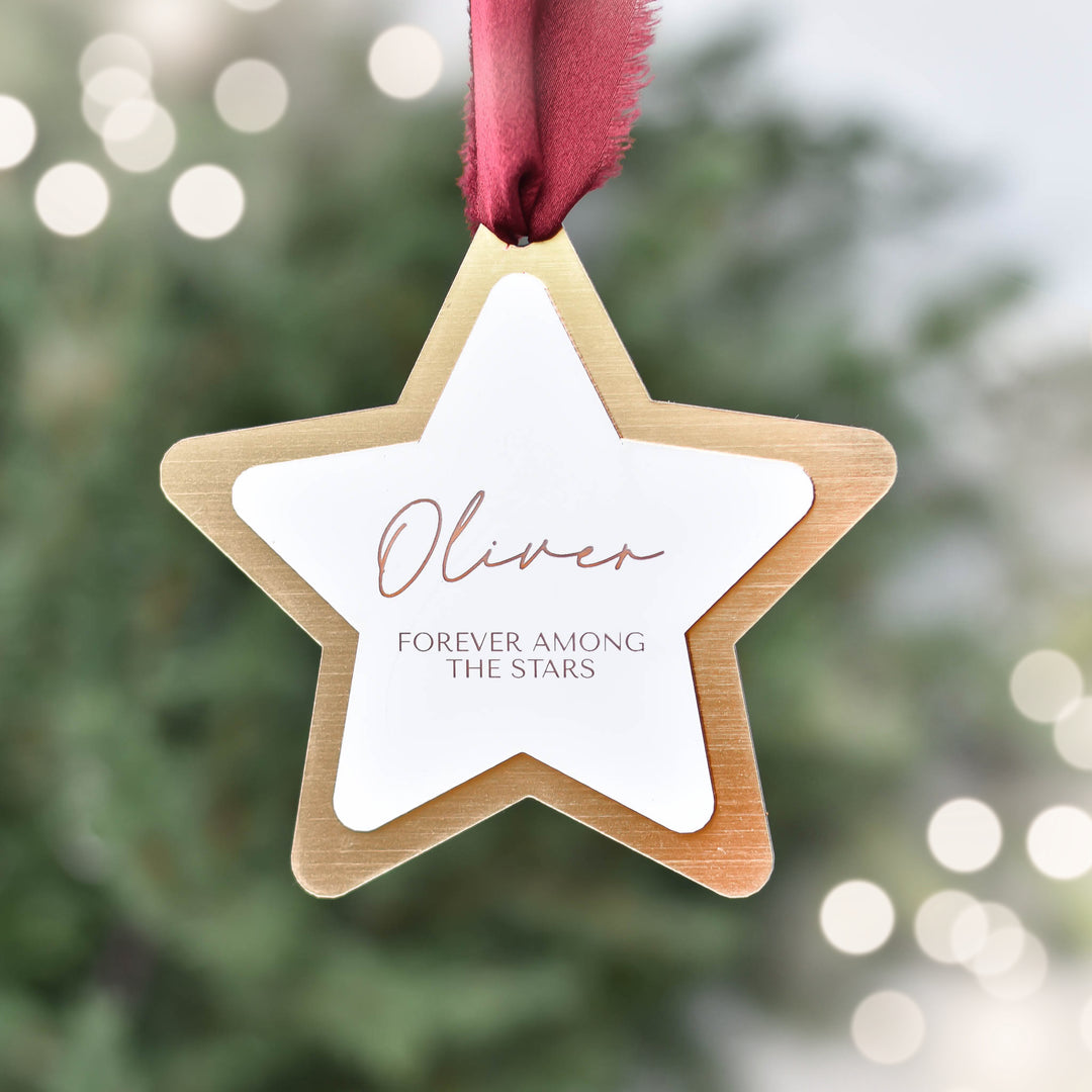 Layered acrylic star ornament in gold and white with the name 'Oliver' and text 'FOREVER AMONG THE STARS'. Red silk ribbon attached.
