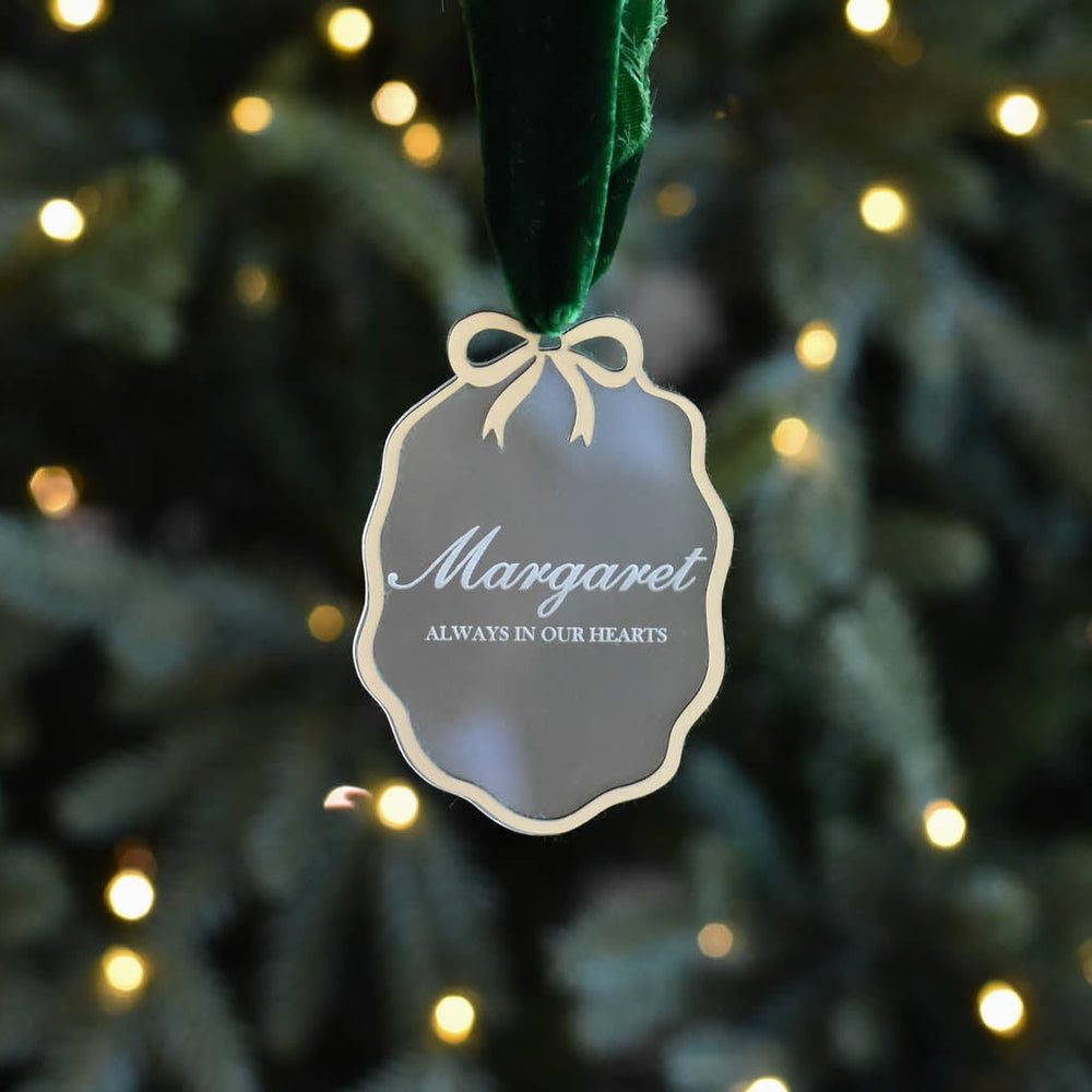 A frosted acrylic ornament with a gold rim and bow at the top, with the words "Margaret, Always in Our Hearts" on the frosted acrylic.