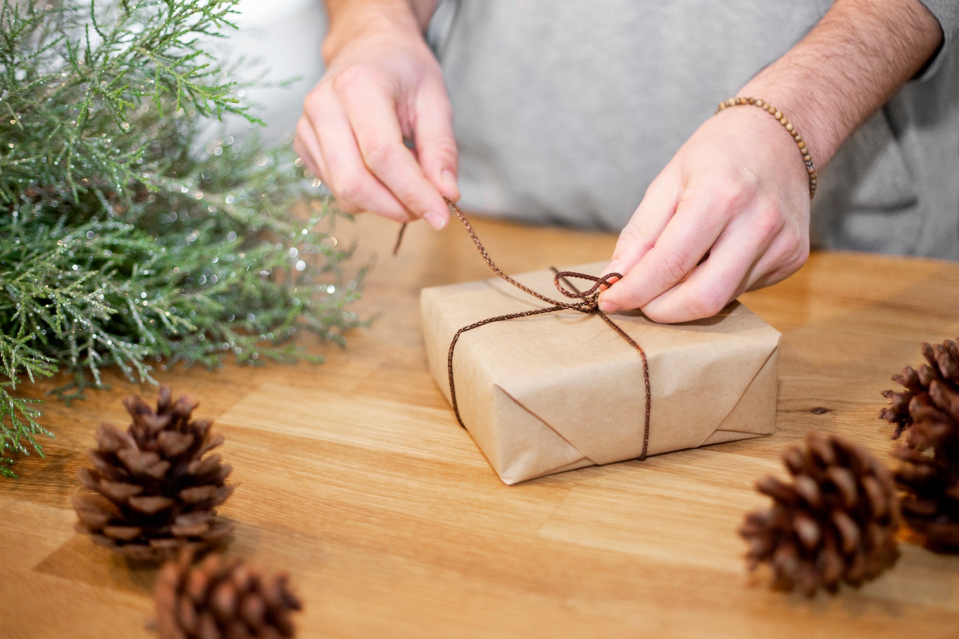 a present wrapped in brown paper being wrapped with a brown bow on a tabletop with pine cones and glittery tree branches