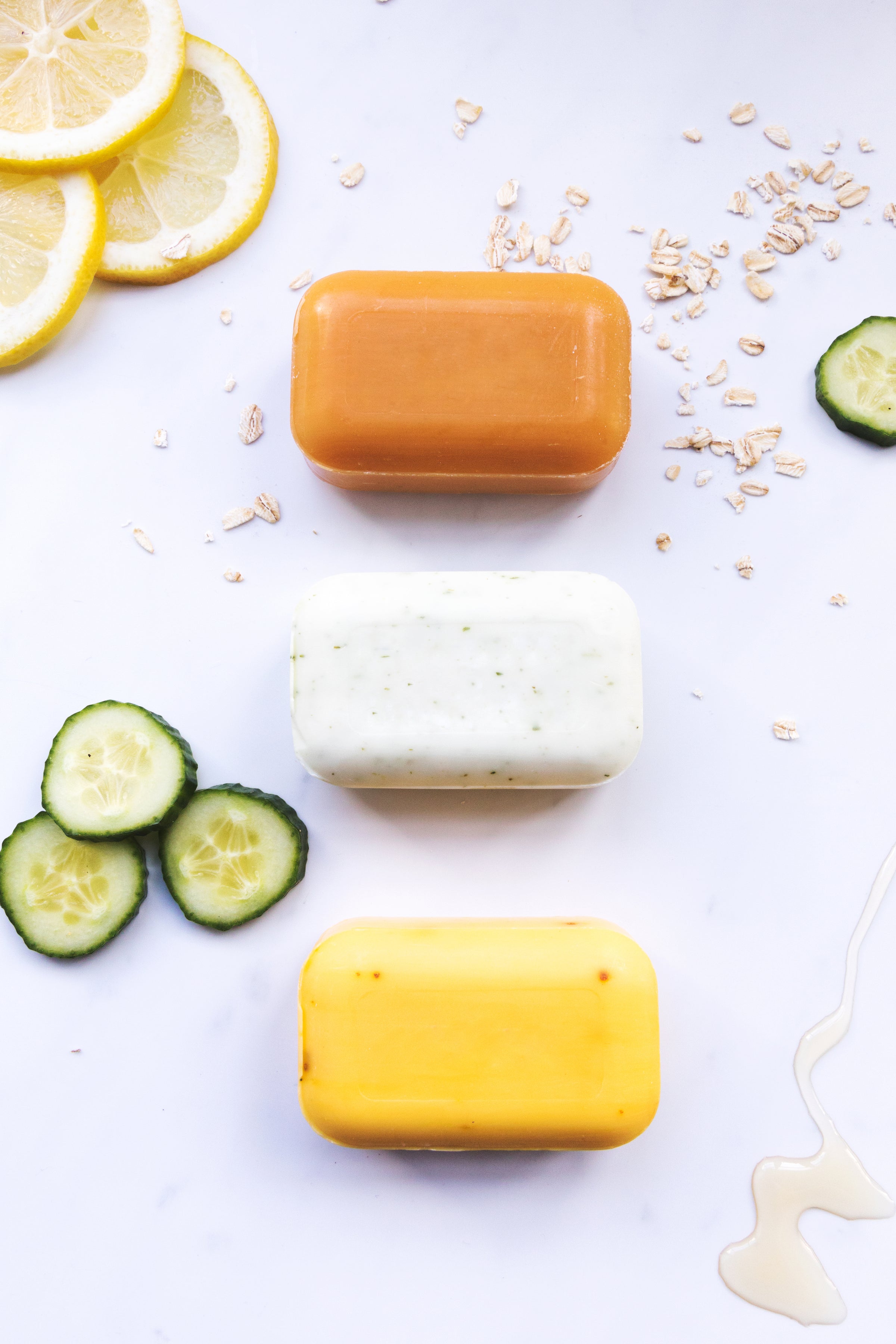 an orange bar of soap, white speckled bar of soap and a yellow bar of soap sitting in a vertical row next to slices of lemon, cucumber and dry oats