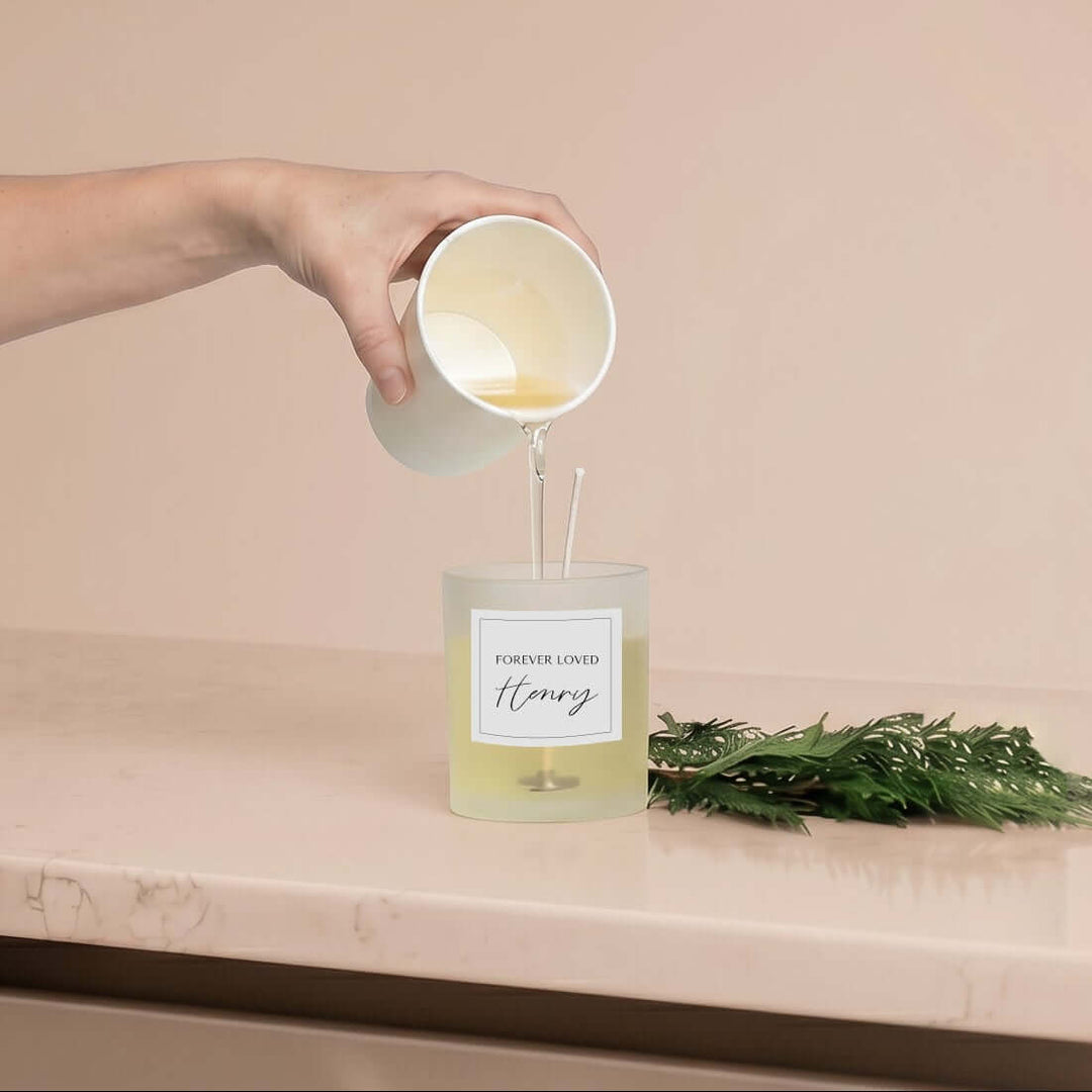 Candle Refill Kit, pouring liquid wax into frosted glass candle labeled 'Forever Loved Henry', green foliage nearby.