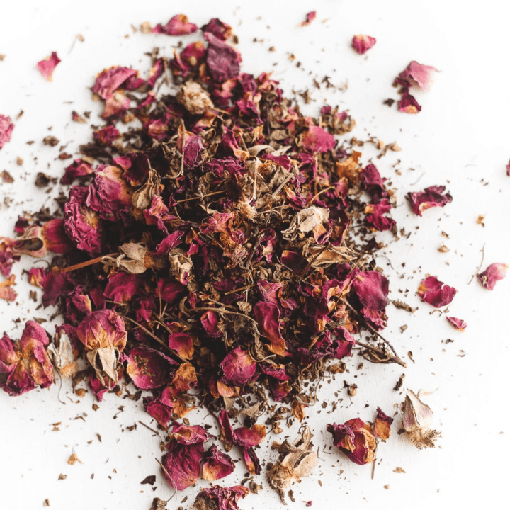 Dried rose petals and herbs in shades of pink and brown, scattered on a white surface. Cup of Love Herbal Tea.