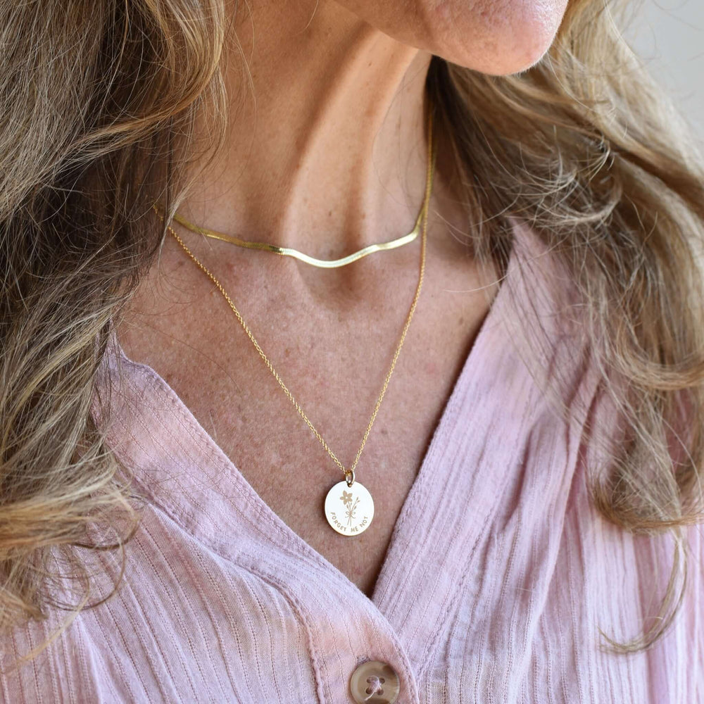 Forget Me Not Necklace in gold on a woman, featuring a floral engraving and delicate chain, layered with a curved gold choker.