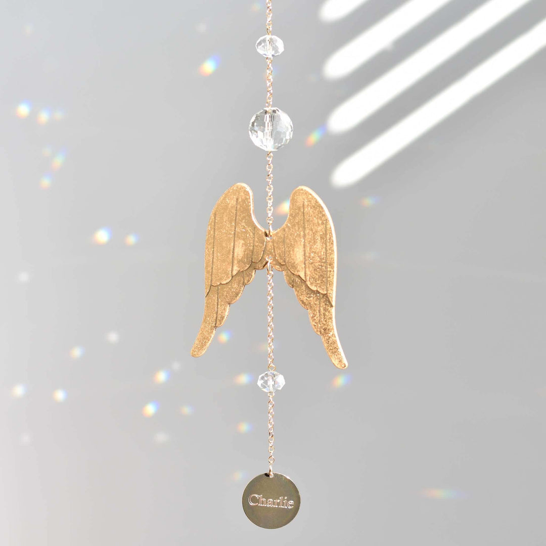 Light of an Angel golden angel wing sun catcher with crystal beads and engraved disc hanging against grey background.