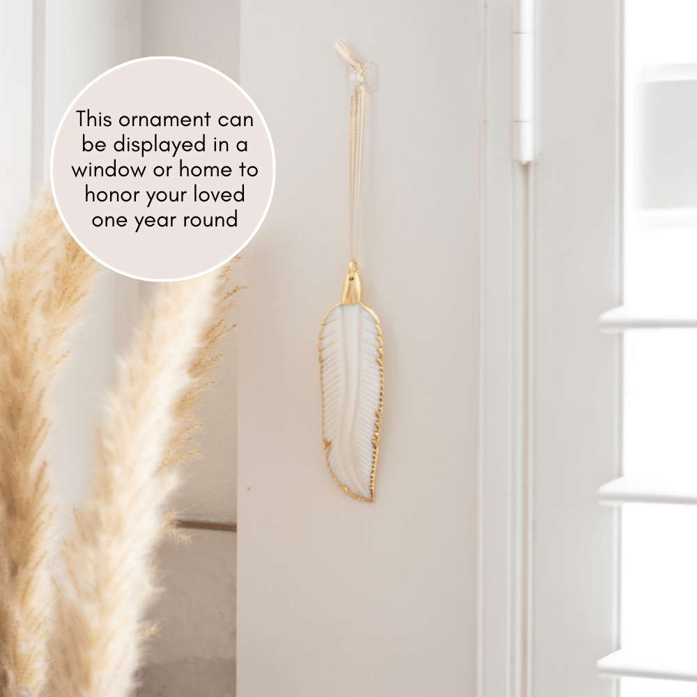 In Memory Feather Ornament, white feather with gold edges, hanging on a wall next to decorative grass.