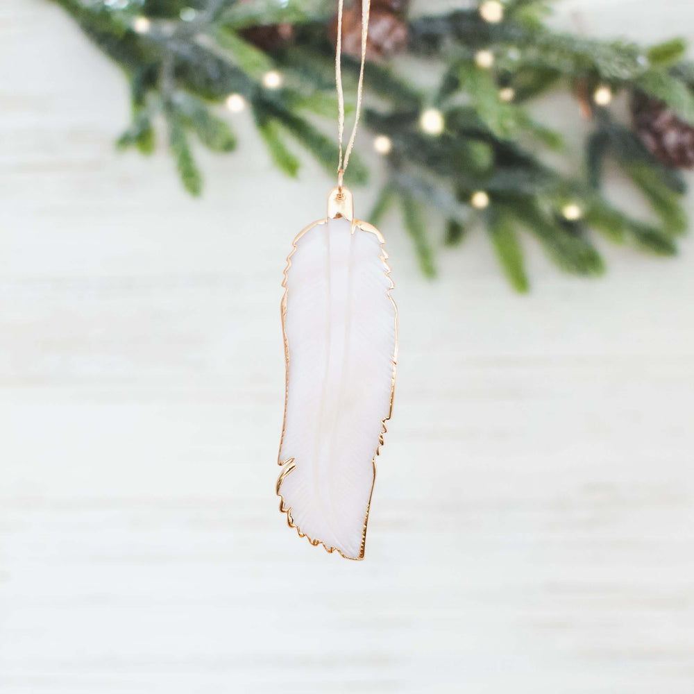 Sound of an Angel pendant shaped like a white feather with gold edging, hanging on a string near green foliage.