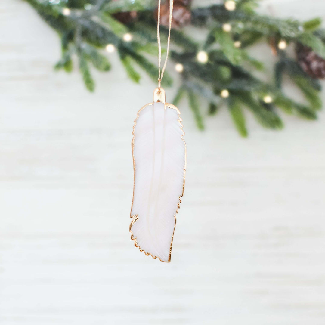 Sound of an Angel pendant shaped like a white feather with gold edging, hanging on a string near green foliage.