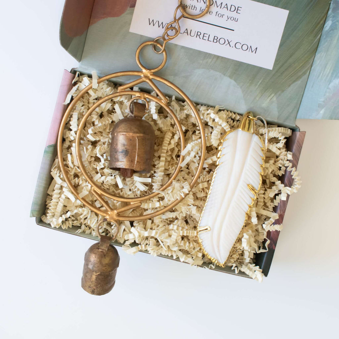 Sound of an Angel gift box with a brass bell and white feather ornament on crinkled paper.