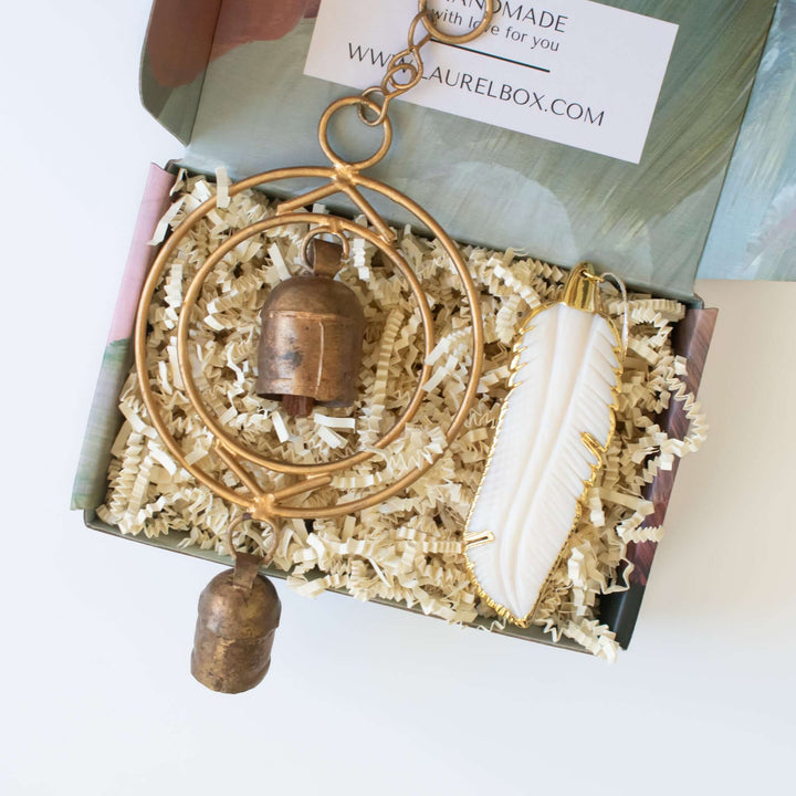 Sound of an Angel gift box with a brass bell and white feather ornament on crinkled paper.