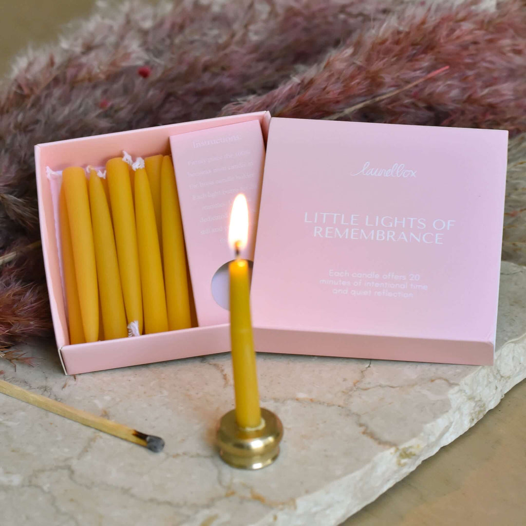 Yellow candle lit in a brass holder next to a pink box labeled "Little Lights of Remembrance" with candles inside.