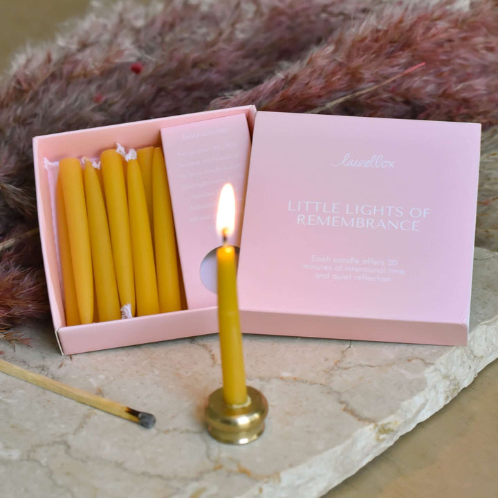 Yellow candle lit in a brass holder next to a pink box labeled "Little Lights of Remembrance" with candles inside.