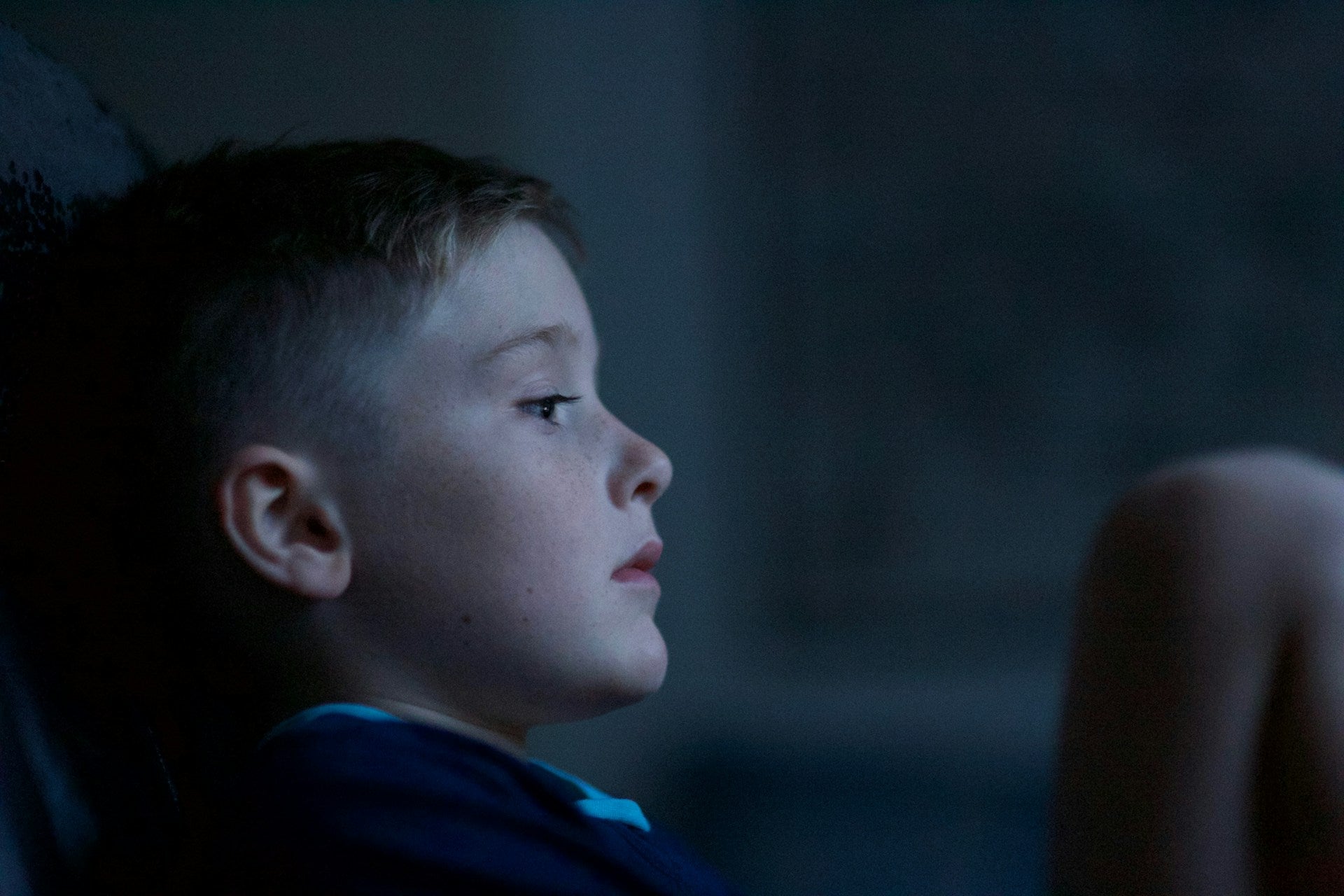 Young boy sitting in a dark room, looking intently at something off-camera.