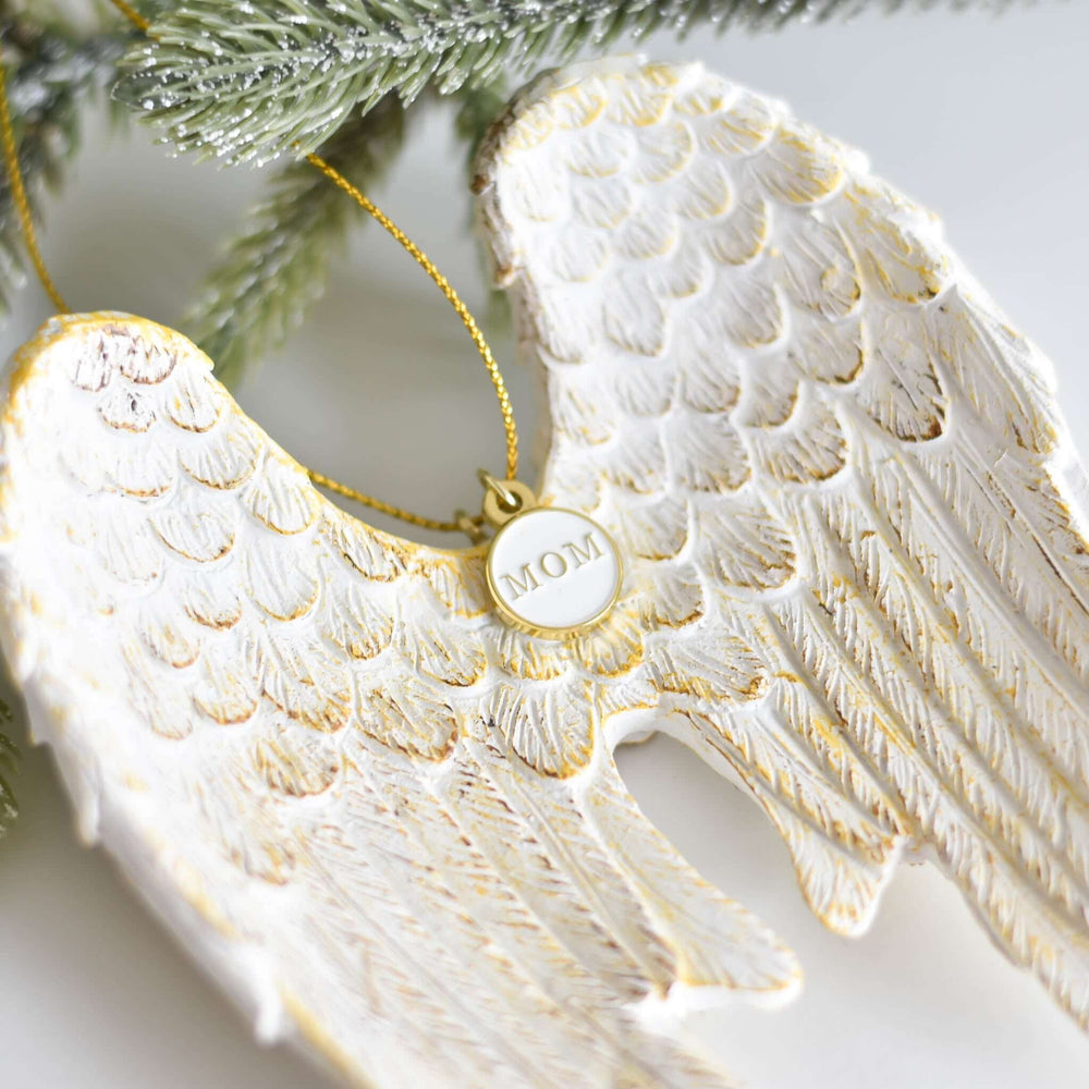 Memorial Angel Wing Ornament in white with gold detailing, featuring a gold 'MOM' tag. Measures 5x4 inches, hanging from greenery.