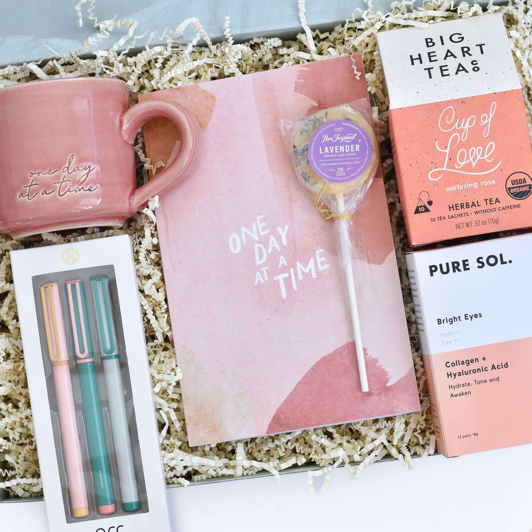 One Day at a Time Gift Box with a pink mug, notepad, tea, lollipop, and pens on shredded paper.