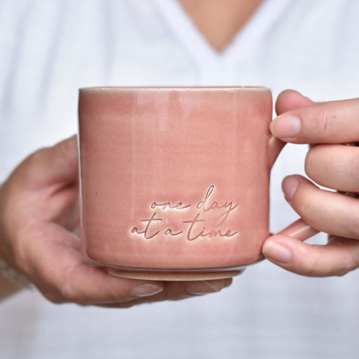 One Day at a Time Mug in soft pink ceramic, featuring embossed text "one day at a time" on the side, held by a hand.