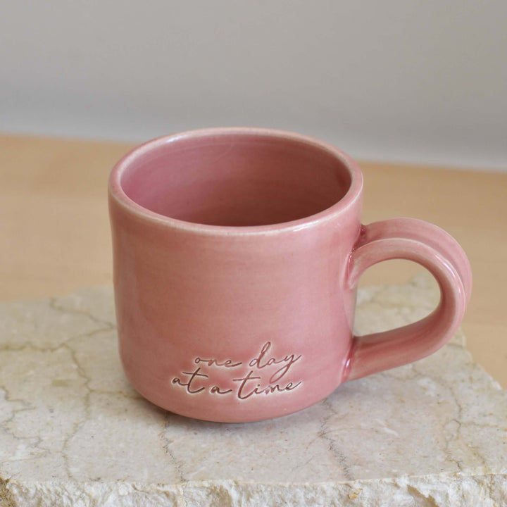 One Day at a Time Mug, hand-thrown pink pottery mug with engraved text on the side, sitting on a stone surface.