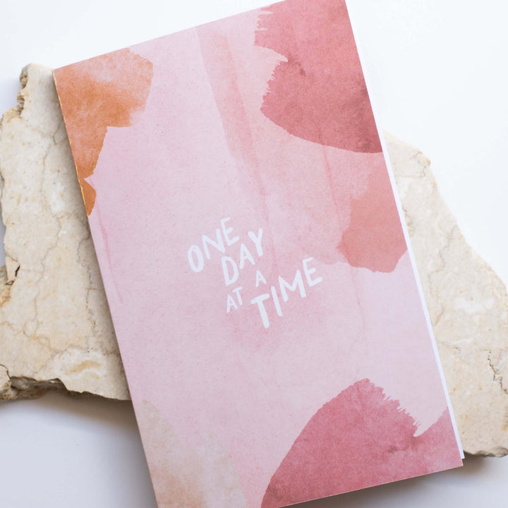 One Day at a Time Journal with pink watercolor cover featuring white text; set on a light stone background.
